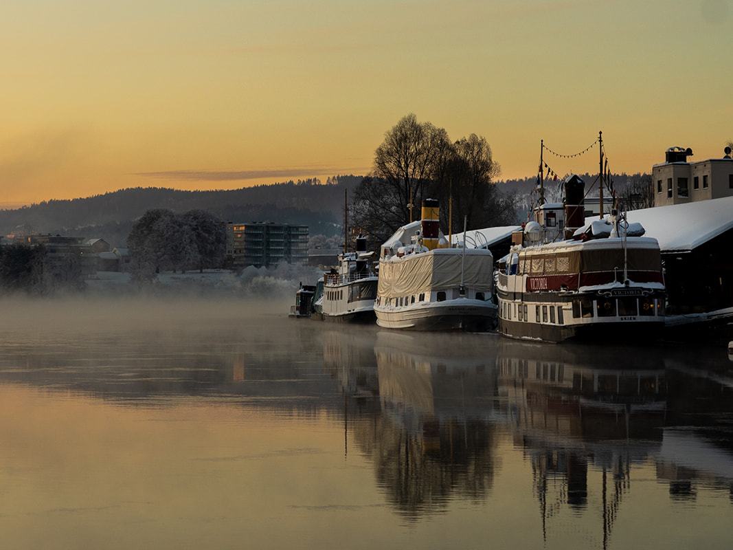 Gamle dampskip ved kaia en tåkete soloppgang, med speilblankt vann og byen i bakgrunnen